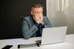 A man in a dark grey suit with a black shirt sitting at a white table with a laptop in front of him. He has his hands over his mouth in frustration.